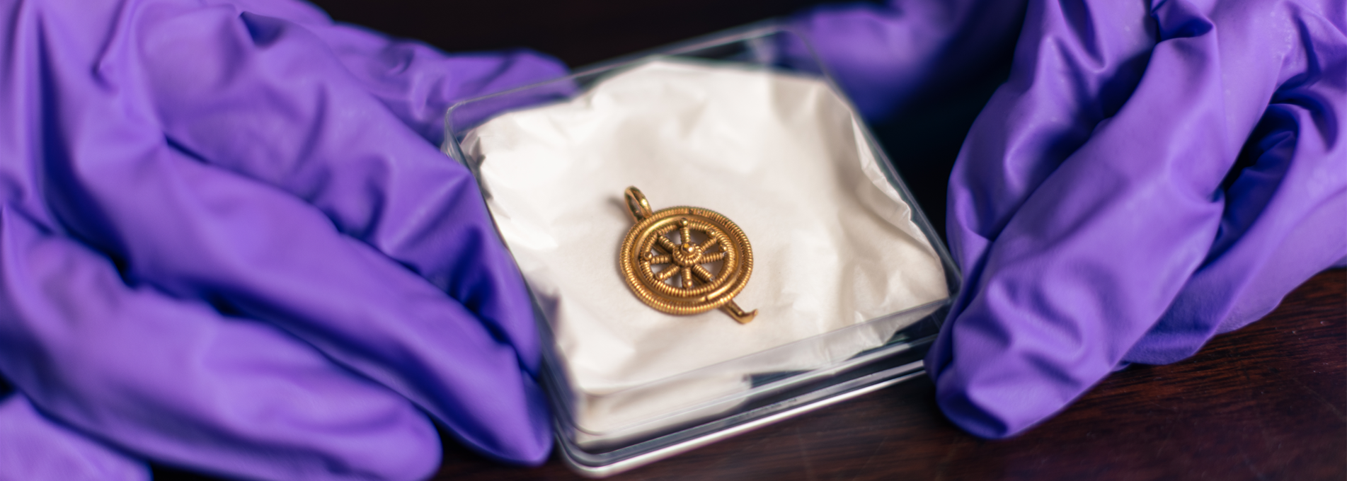 Small gold pendant in a box, being held up by gloved hands