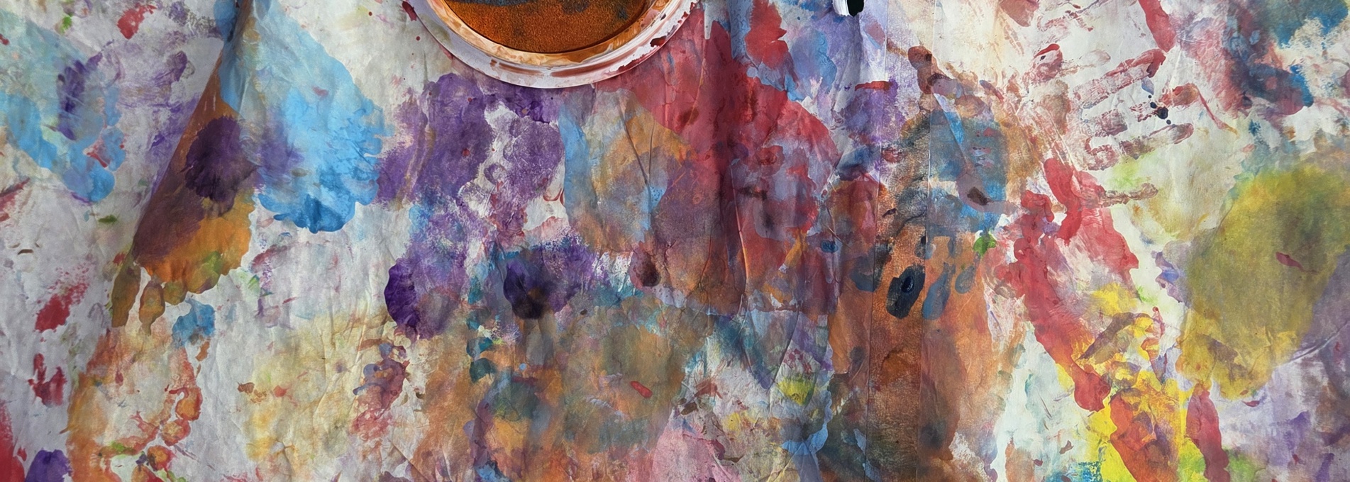 Overhead view of a large sheet of paper covered with colorful paint splatters, handprints, footprints, and brush marks in shades of blue, purple, red, yellow, green, and orange. Several open paint containers and sponge paint pads sit near the top of the image, suggesting a children's arts and crafts activity. The surface is messy and vibrant, filled with layered paint impressions and creative marks.