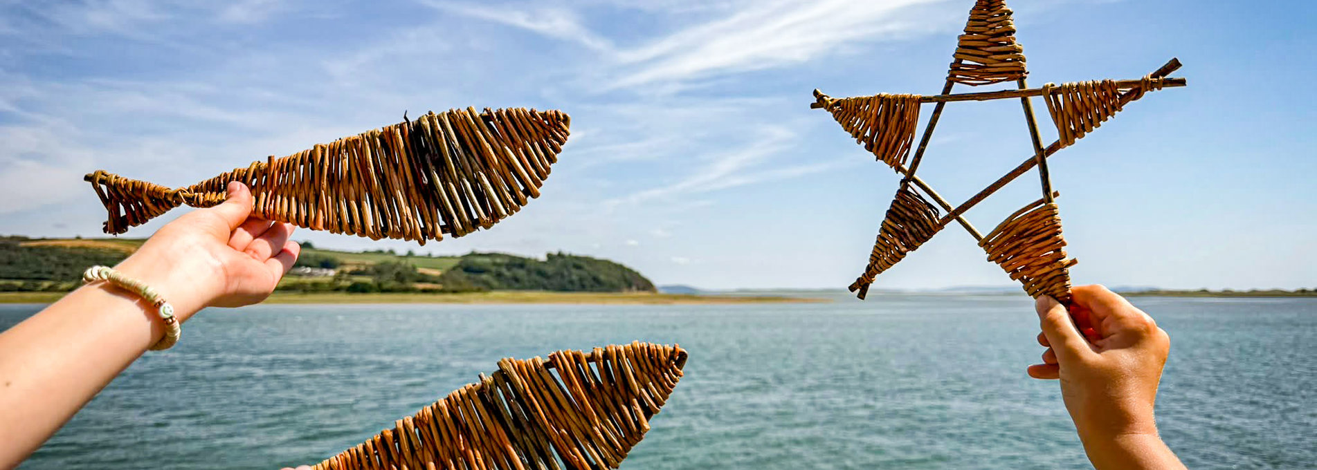 Hands holding handmade willow fish and star-shaped woven decorations against a coastal backdrop, with estuary waters and shoreline visible in the distance