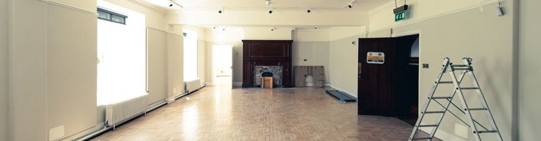 Wide open room with sunlight streaming through two windows on the left wall. A fireplace is at the rear of the room and an open door near a ladder are on the right.